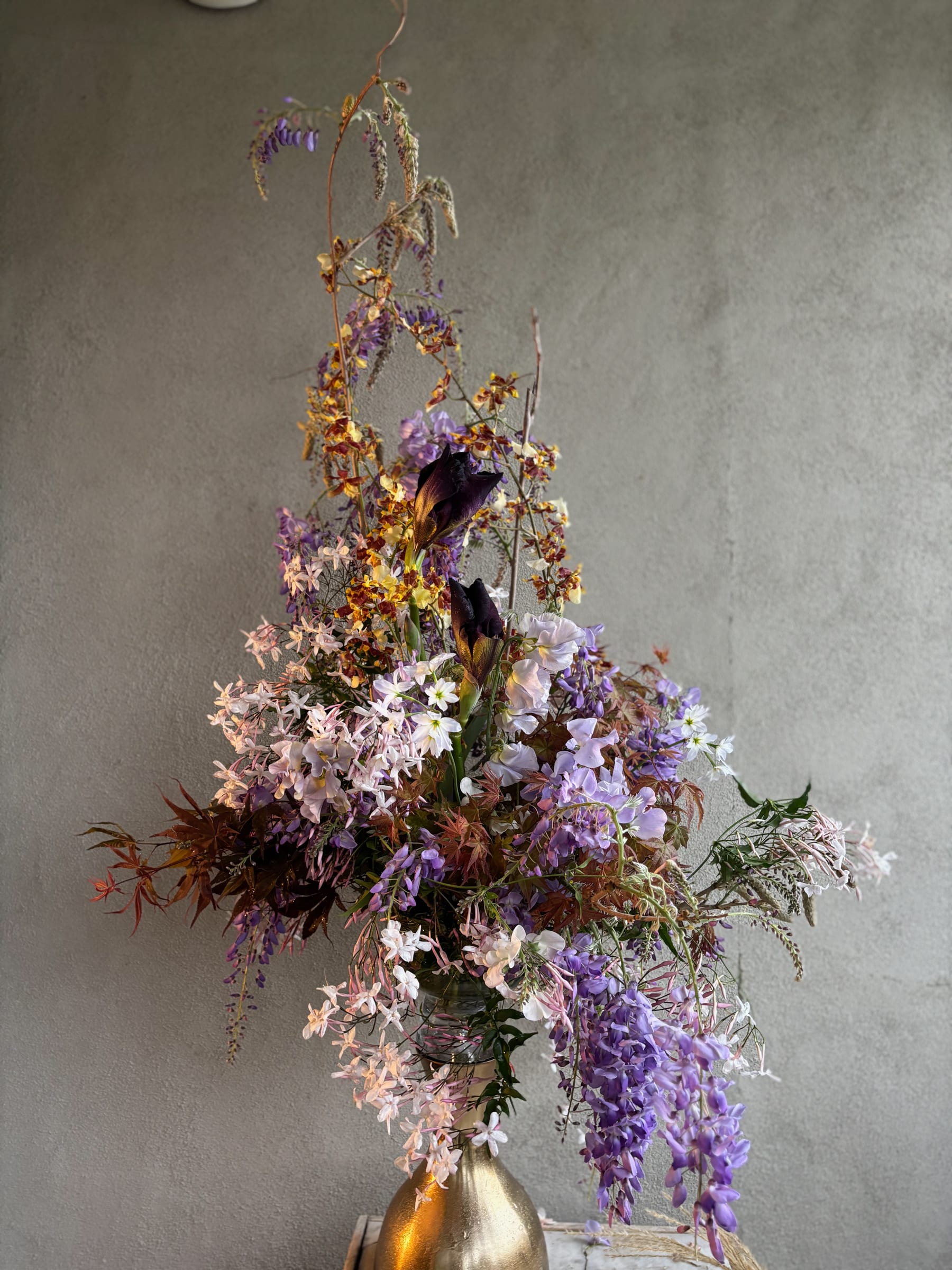 Tall sculptural arrangement of wisteria, oncidium orchids, and iris in a brass vessel